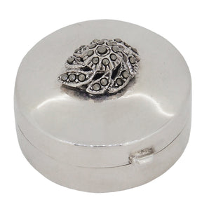 A modern, white metal, circular pill box with a marcasite set floral motif on the hinged lid.