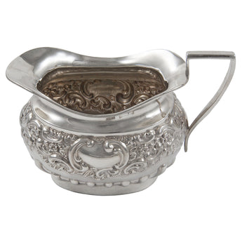 An Edwardian, silver cream jug with embossed floral, foliate & scroll decoration & a vacant cartouche.