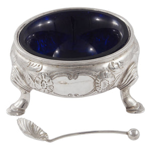 A Victorian, silver, circular open salt with floral decoration, vacant cartouche, on three feet with a silver spoon & blue glass liner.
